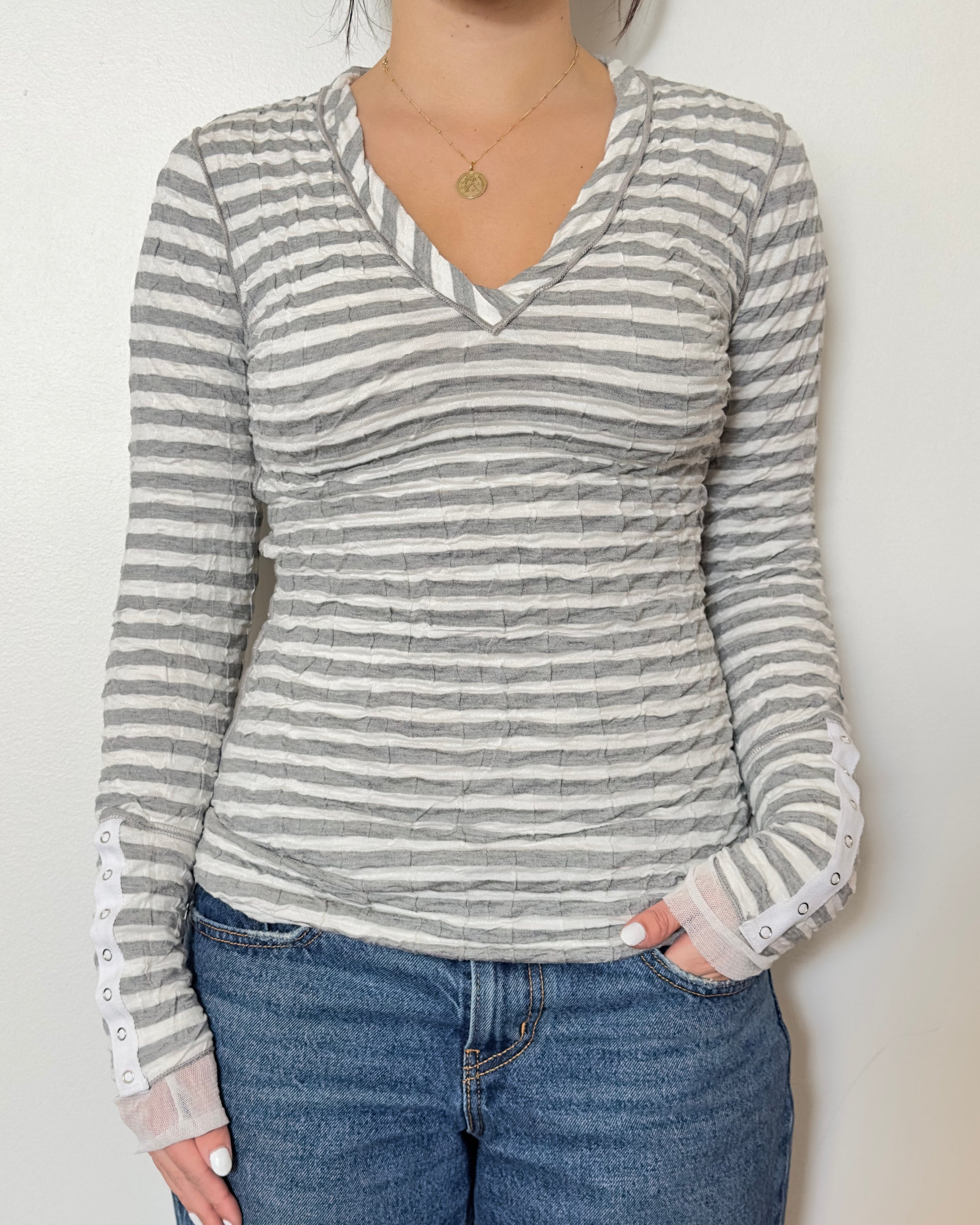 Female model wearing a scrunched striped long sleeve top in a grey and white color with a v neck cut and buttons on the sleeves