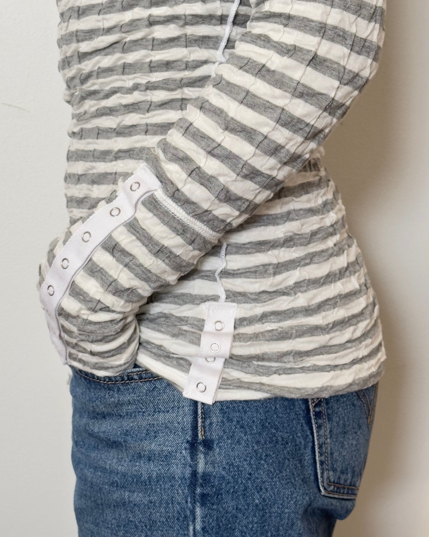 Female model wearing a scrunched striped long sleeve top in a grey and white color with a v neck cut and buttons on the sleeves