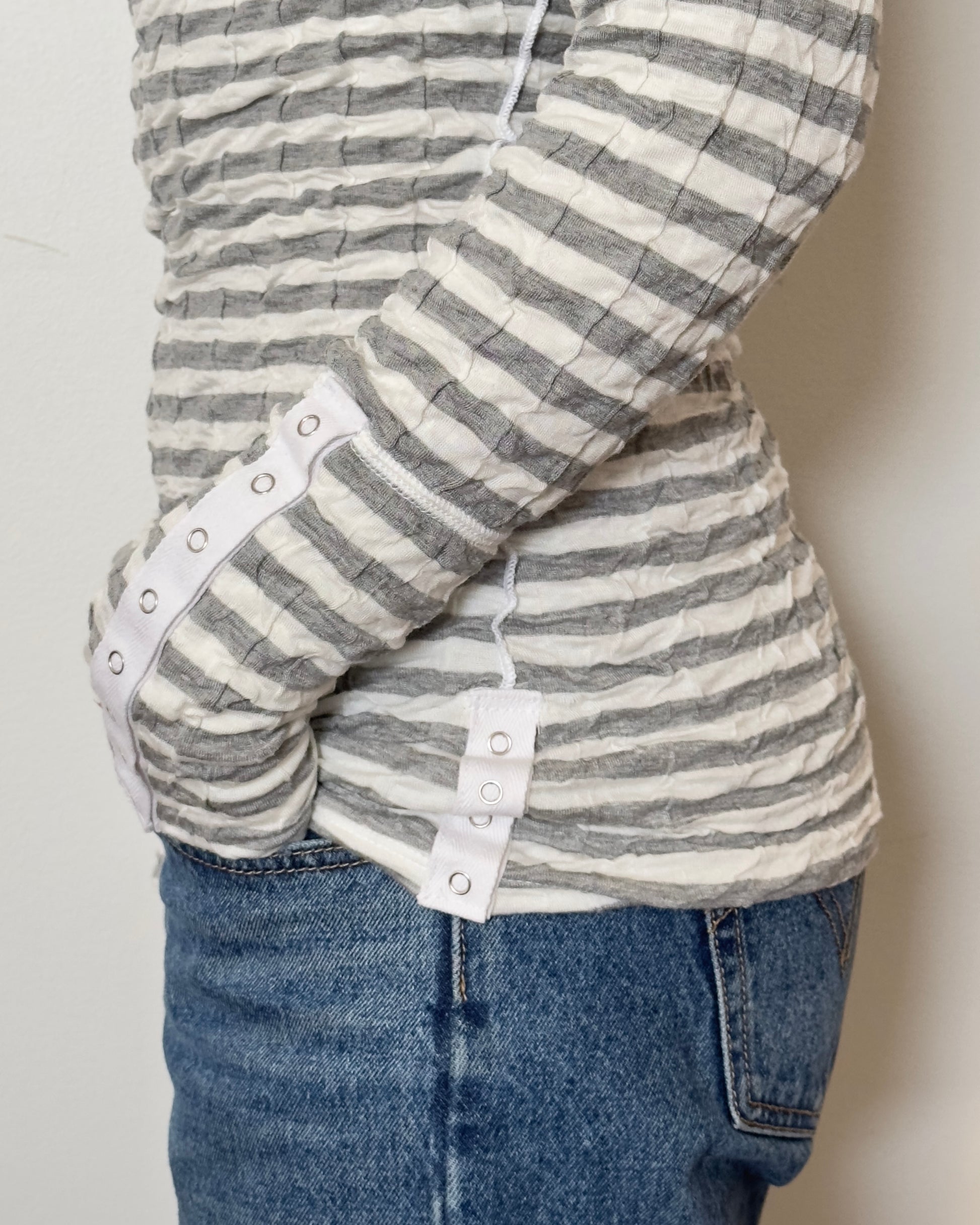 Female model wearing a scrunched striped long sleeve top in a grey and white color with a v neck cut and buttons on the sleeves