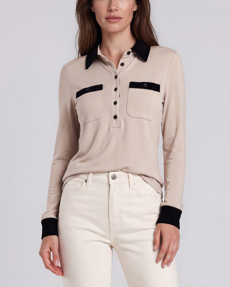 A woman stands against a light gray studio background wearing an oatmeal nude long-sleeve polo shirt with a black collar and chest pocket trim, white jeans. contrast collar polo - pete collection