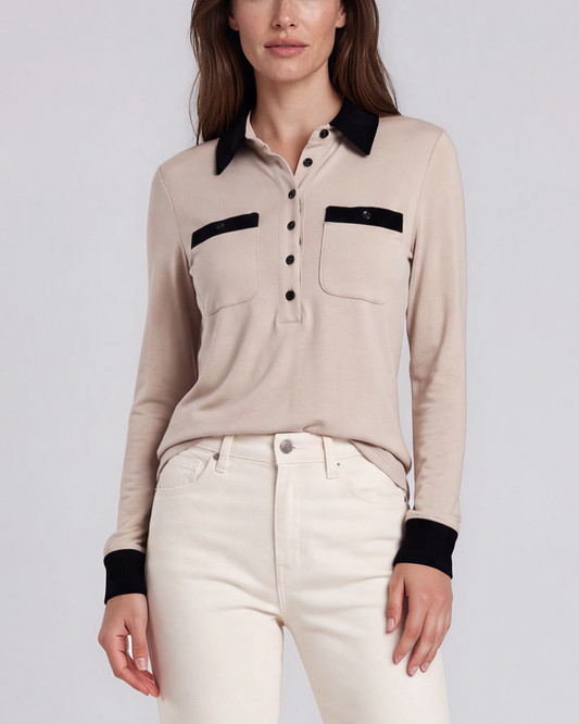 A woman stands against a light gray studio background wearing an oatmeal nude long-sleeve polo shirt with a black collar and chest pocket trim, white jeans. contrast collar polo - pete collection