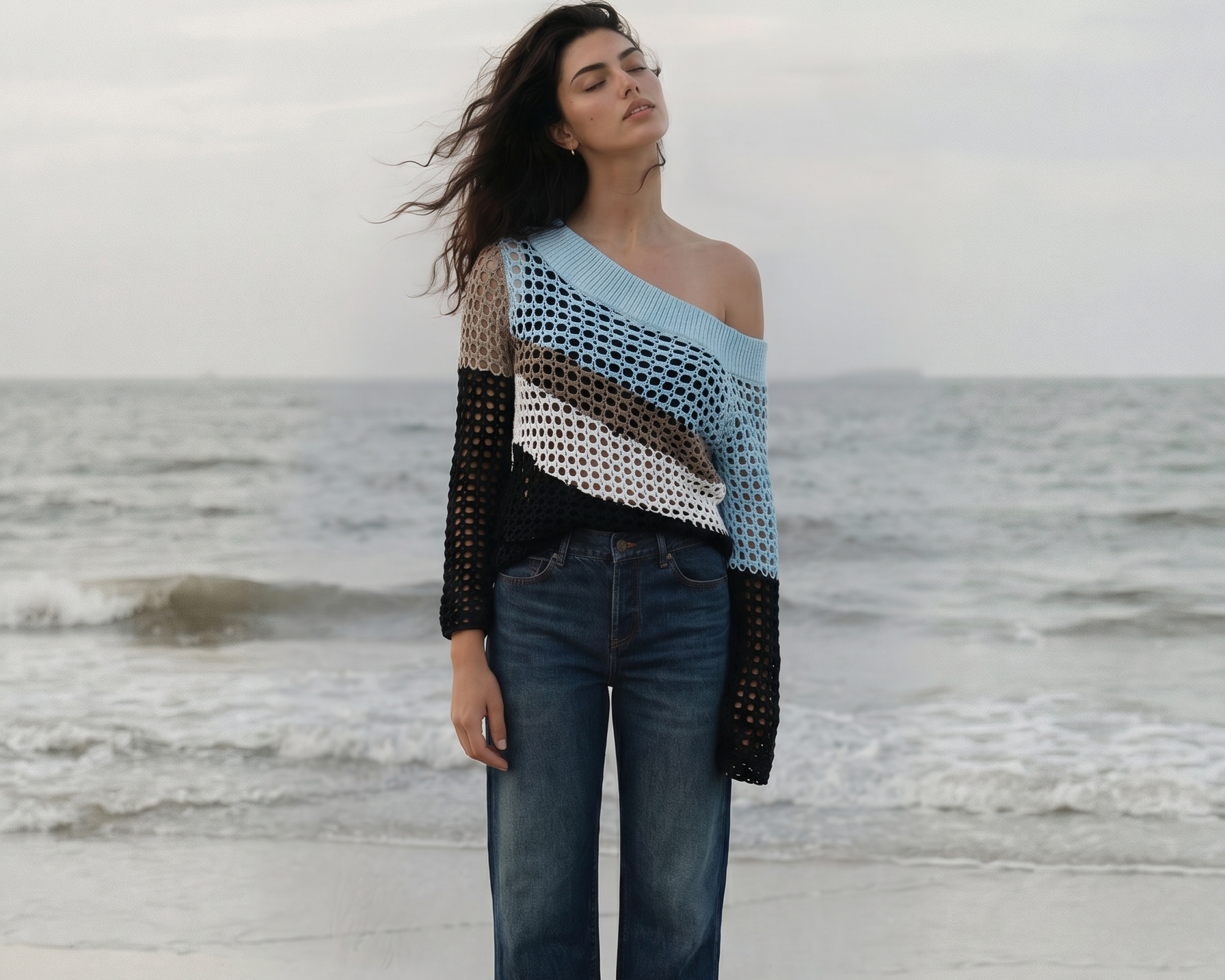 A female model standing on a chair by the ocean, wearing a patterned sweater and jeans. wearing a one-shoulder asymmetrical crochet top in crystal blue, bark (brown), coconut milk (white), and black. Textured knit. - pete collection